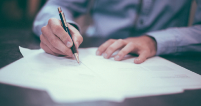 A person in grey business attire is signing a paper document.