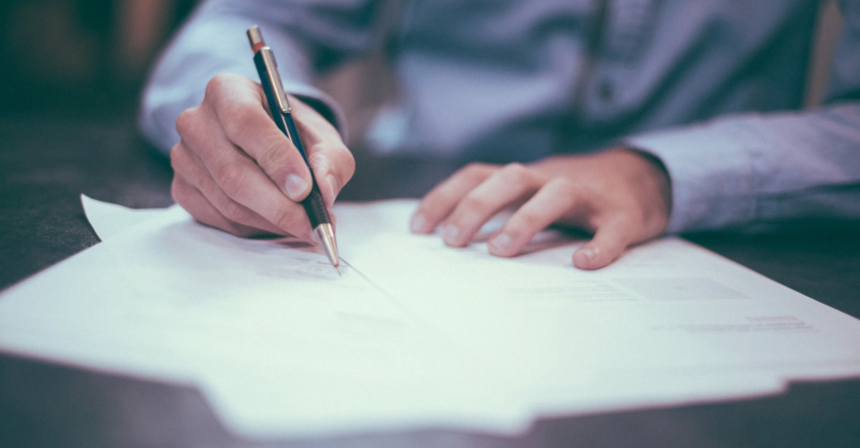 A person in grey business attire is signing a paper document.