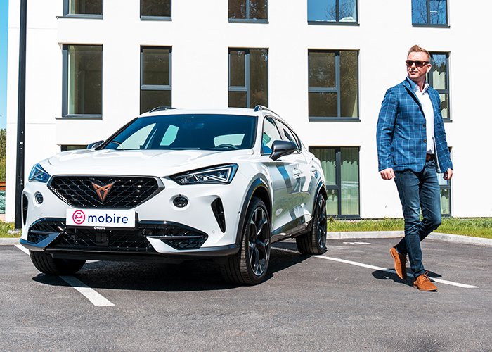 A man standing next to a white car with a Mobire logo on a number plate, with white building in the background.