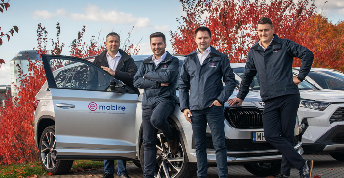 Four men in Mobire jackets standing next to a silver car with a Mobire logo, with red autumn trees in the background.