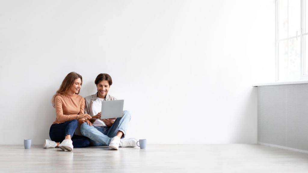 A pair of people sitting on the floor in an empty apartment, discussing something that is seen on a laptop screen.